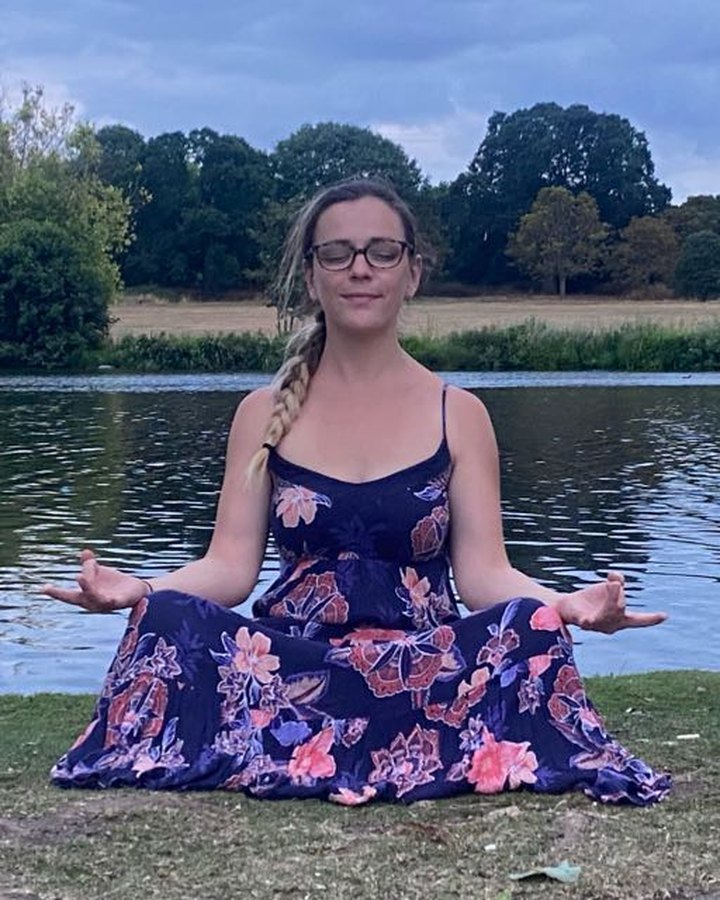 Seated cross-legged in lotus pose beside a lake at dusk